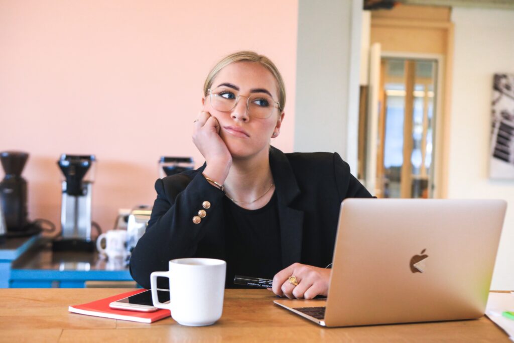 Main image for the survey fatigue blog. Bored woman at a desk with a MacBook in front of her, staring into the distance resting her chin on her hand. Image by Magnet.Me on Unsplash.