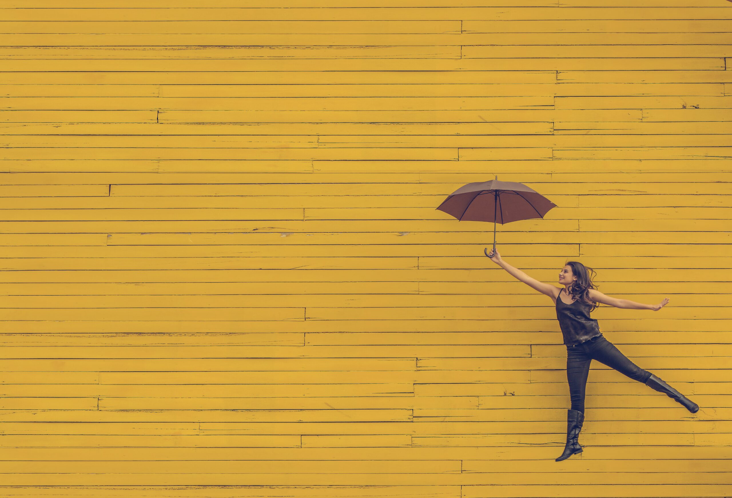 A woman is floating in the air, holding a full umbrella up like Mary Poppins, pictured against a yellow brick wall, grinning and surreal. Represents an employee engagement strategy that boosts morale. Image by Edu Lauton via Unsplash.