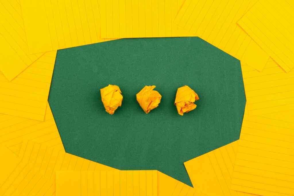 Picture of a speech bubble created using negative space – there is a green background carved into the shape of a bubble by a lot of yellow lined pieces of card. Inside the speech bubble, there are three scrumpled pieces of this yellow card which form an ellipsis. Photo by Volodymyr Hryshchenko on Unsplash