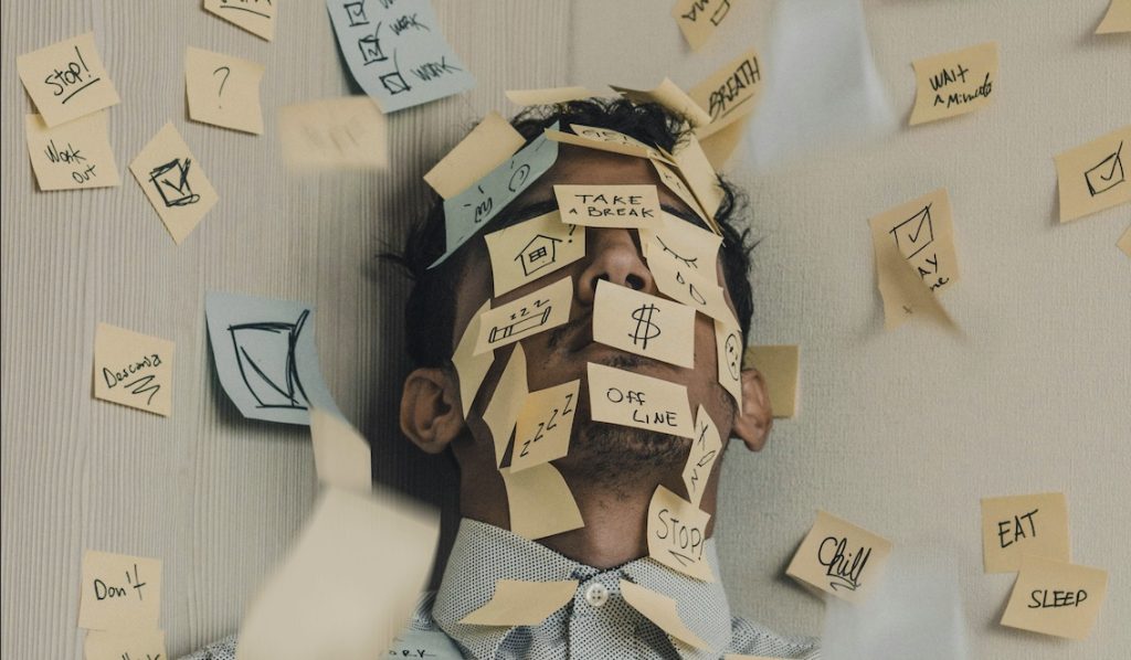 Man squished into a corner with sticky notes covering his face and body saying things like 'Take a break', 'Off line' and 'Stop!'. Image by Luis Villasmil via Unsplash. Thanks Luis.